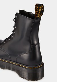 Botte noire en cuir à lacets avec semelle épaisse rainurée, coutures jaunes, et languette noire et jaune à l'arrière.