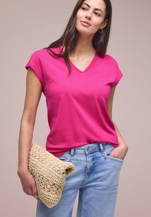 Femme portant un t-shirt rose vif à col en V et un jean bleu clair, tenant une pochette beige tissée dans la main gauche, main droite dans la poche.