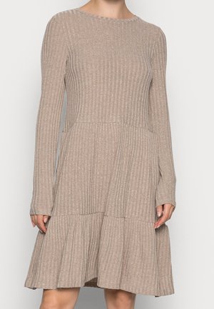 Beige geribbelde gebreide jurk met lange mouwen, getailleerde taille en geplooide uitlopende rok, gedragen door een staande persoon tegen een effen achtergrond.