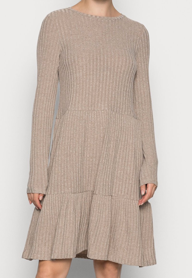 Robe en tricot côtelé beige avec manches longues, taille cintrée et jupe évasée à étages, portée par une personne debout sur un fond uni.