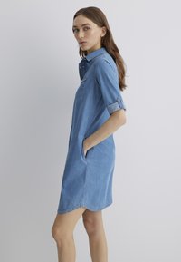 Denim shirt dress in light blue, featuring a collar, rolled-up sleeves, side pockets, and a slightly curved hemline.