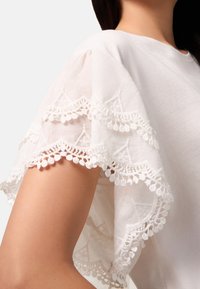 Blusa bianca con maniche corte e svasate, caratterizzata da una preziosa rifinitura in pizzo e dettagli con pom-pom. Tessuto morbido, vestibilità comoda e leggera texture.