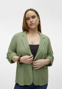 Blazer vert clair en tissu doux, avec un col cranté, des manches retroussées et deux poches avant. Porté par-dessus un haut noir.
