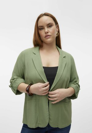 Blazer vert clair en tissu doux, avec un col cranté, des manches retroussées et deux poches avant. Porté par-dessus un haut noir.