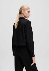 Zwarte cropped shirt met lange mouwen en een kraag met knopen, met een gladde textuur en strakke lijnen. De broek is ook zwart en heeft een getailleerde pasvorm.