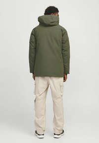 Chaqueta verde oliva con capucha de textura suave, combinada con pantalones cargo de color beige claro que presentan múltiples bolsillos, vista desde la parte posterior.