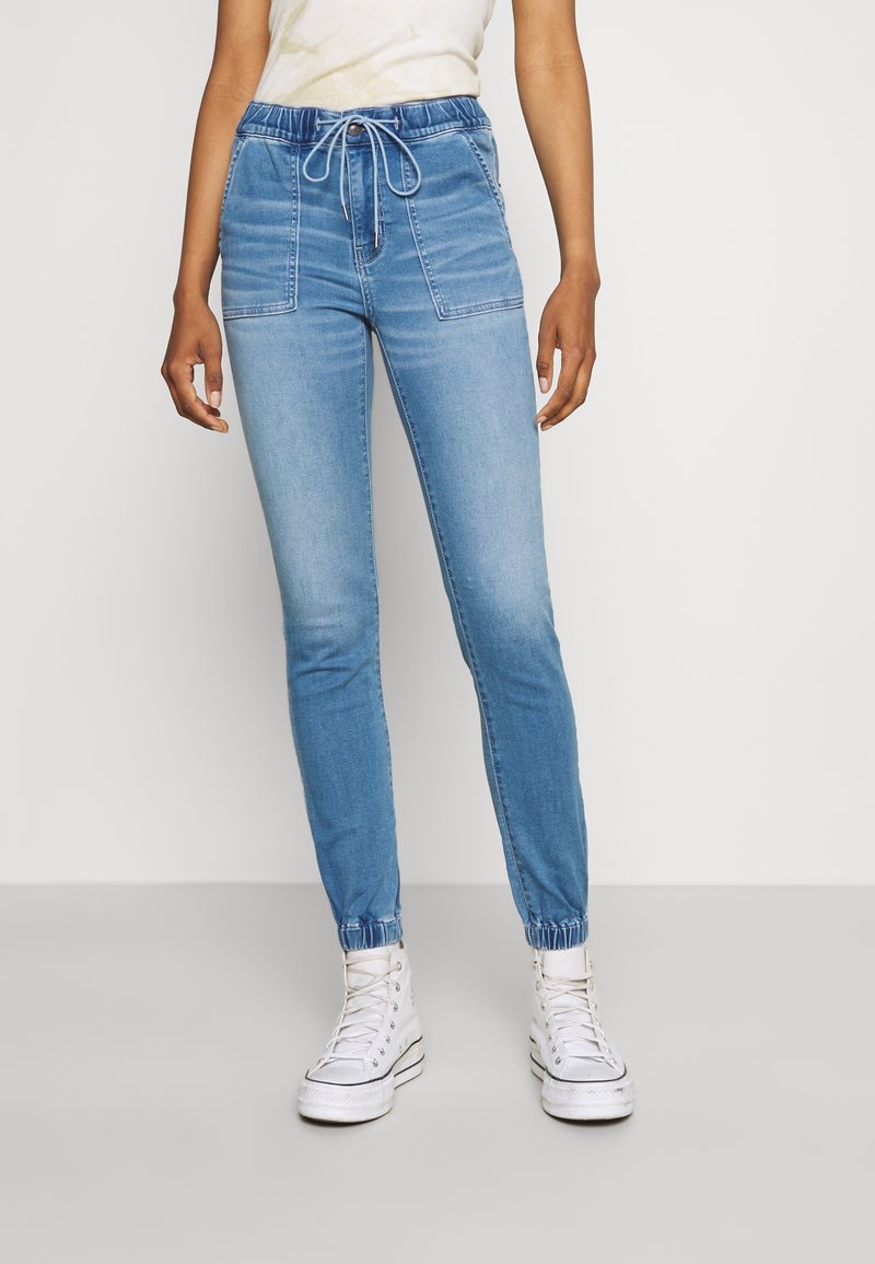 Personne portant un pantalon jogger en denim bleu clair avec poches avant et des baskets montantes blanches, debout devant un fond uni.