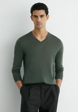 Oliwkowozielony sweter z dekoltem w serek, lekki, dopasowany fason z długimi rękawami i prążkowanymi mankietami, zestawiony z ciemnymi spodniami. Teksturowana mieszanka bawełny.