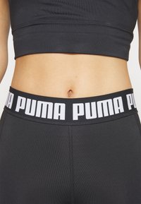 Mustat urheilulegginsit, joissa on leveä vyötärönauha ja valkoiset "PUMA"-kirjaimet. Materiaali on sileä ja tiukka, korostaen virtaviivaista muotoa.