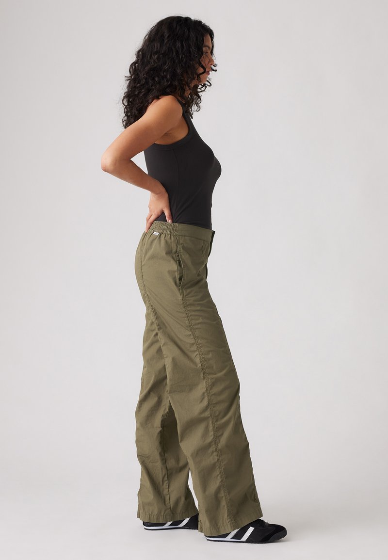 Pantalons cargo à taille élastique en vert olive, fabriqués en tissu léger, avec des poches latérales et une coupe décontractée à jambes larges. Chaussures noires avec des rayures blanches.