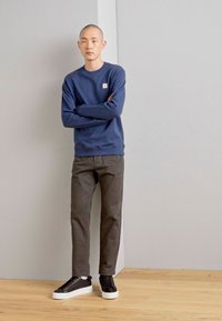 Hanorac bleumarin cu guler rotund, chinos gri și adidași negri cu talpă albă. Modelul stă drept în fața unui fundal simplu.