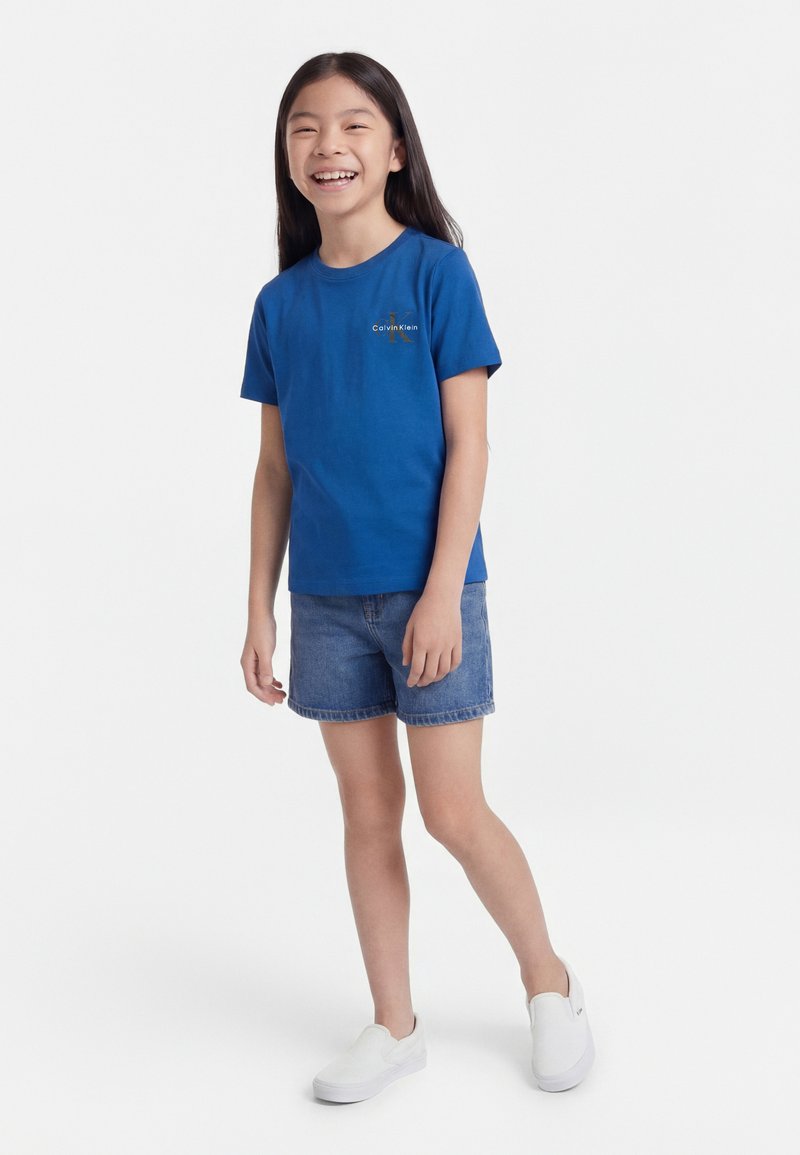 Naeratav tüdruk, kes seisab sinises Calvin Kleini t-särgis, teksapükste lühikestes lühikestes pükstes ja valgetes libisemisjalanõudes tavalise valge tausta ees.