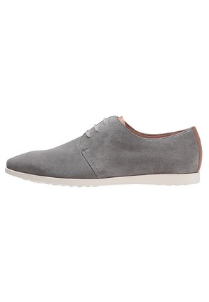 Grå ruskindssko med spids snude, lysfarvet gummisål, minimalistisk snøredesign og brun læderdetalje ved hælen.