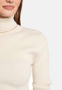 Col roulé crème côtelé au design ajusté et à la texture lisse. Doté d'un tissu léger et d'un subtil accent avec chaîne dorée.