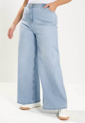 Person trägt hellblaue Jeans mit weitem Bein und weiße Turnschuhe, eine Hand in der Tasche, steht vor einem schlichten weißen Hintergrund.