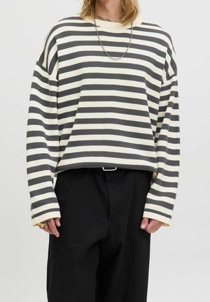 Person trägt ein schwarz-weiß horizontal gestreiftes Langarmshirt, schwarze Hose, eine silberne Kettenhalskette und Ringe an den Fingern.
