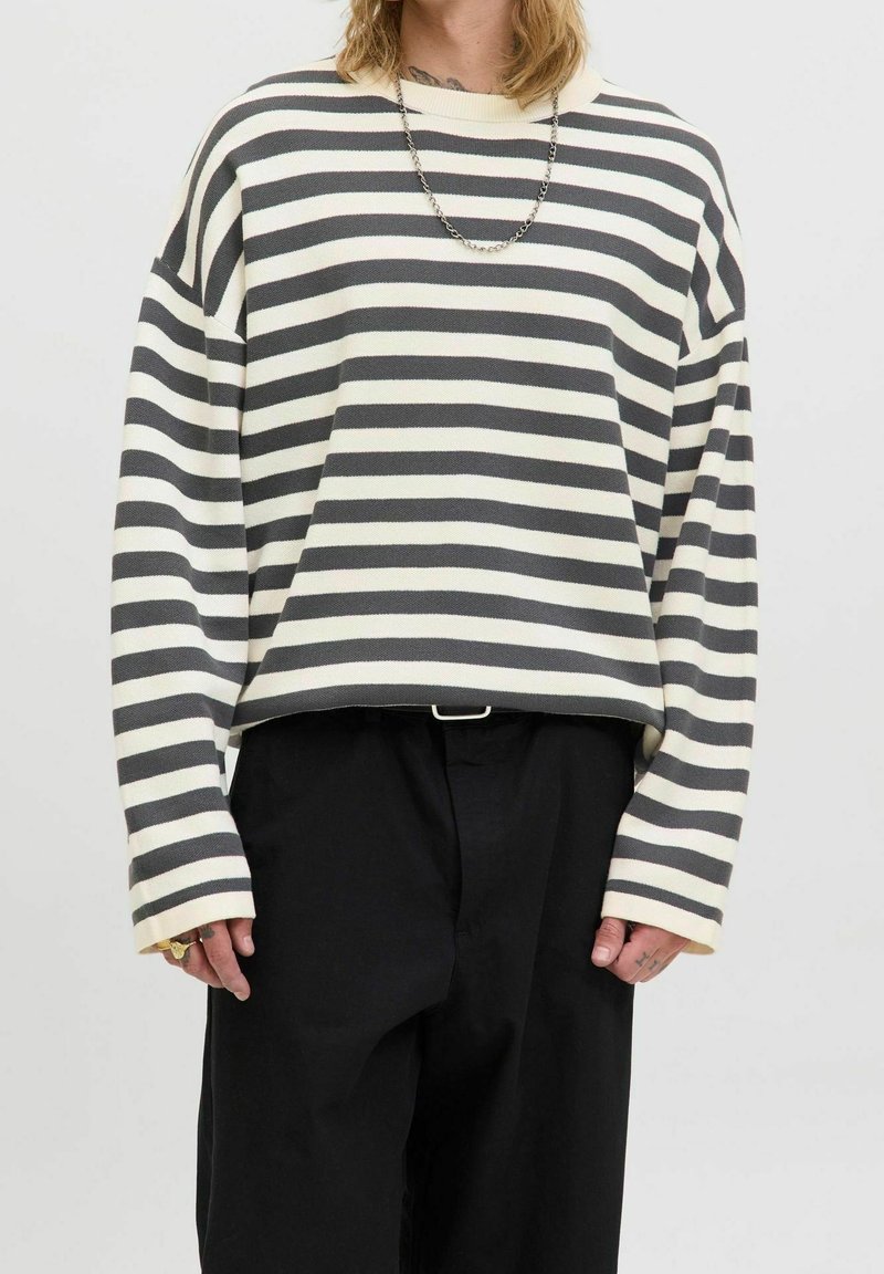 Personne portant un t-shirt à manches longues rayé horizontalement noir et blanc, un pantalon noir, un collier chaîne en argent et des bagues aux doigts.