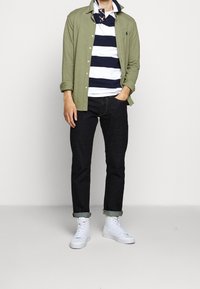 Muž oblečený v olivově zelené košili přes námořnickou a bílou pruhovanou polokošili, tmavých zkrácených džínech a bílých vysokých teniskách stojící proti jednobarevnému pozadí.