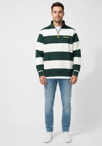 Hombre vestido con un pulóver a rayas horizontales verdes y blancas con media cremallera, jeans de mezclilla azul claros y zapatillas blancas, de pie contra un fondo liso.