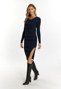 Marineblaues geripptes Kleid mit langen Ärmeln, versehen mit silbernen Nieten und seitlichen Schlitzen. Getragen mit kniehohen schwarzen Absatzstiefeln.