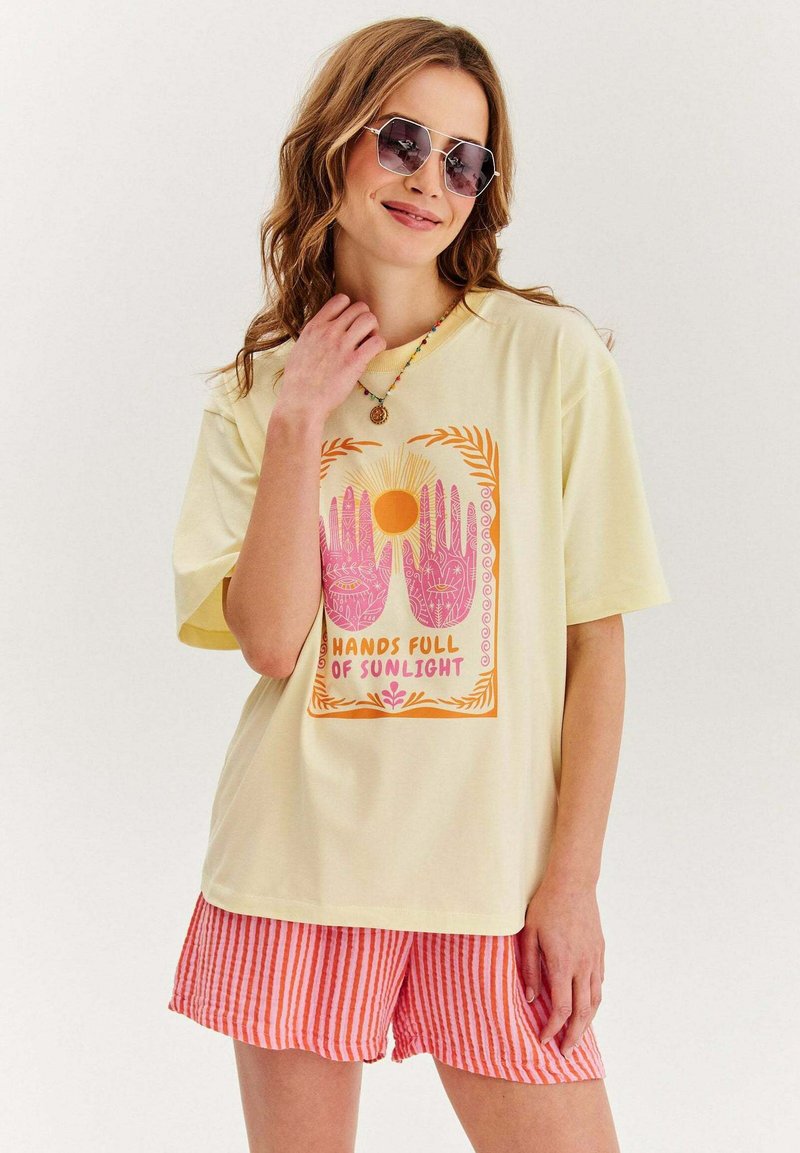Světle žluté oversized tričko s barevným grafickým potiskem rukou a slunce. V kombinaci s šortkami červeno-bílého pruhu, z bavlněného směsového materiálu.