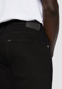 Acercamiento al bolsillo de unos pantalones Lee negros y la etiqueta en la cintura, llevados con una camisa blanca en la parte inferior del torso y el brazo de una persona de piel oscura.