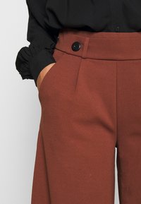 Široké kalhoty cihlově červené barvy z texturované látky, s vysokým pasem, předními kapsami a černým knoflíkovým detailem.