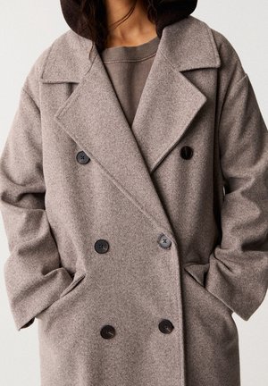 Persoon draagt een taupe double-breasted wollen jas met brede revers en handen in de zakken over een bijpassende trui.