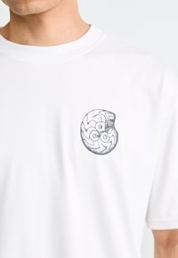 Person wearing white T-shirt with small black abstract circular design and letters "O," "C," "G," and "A" on the chest.
