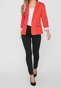 Rode blazer met schoudervulling, sluiting met één knoop en twee voorzakken, gecombineerd met zwarte strakke broek en zwarte hakken. Witte top eronder.