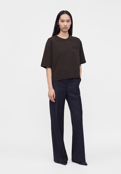 Femme aux longs cheveux noirs lisses portant une chemise noire ample à manches courtes et un pantalon large et sombre, debout devant un fond blanc.