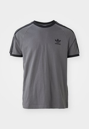 T-shirt gris à manches courtes avec des accents noirs. Il présente un col rond, trois rayures noires sur les épaules et un petit logo noir sur la poitrine.