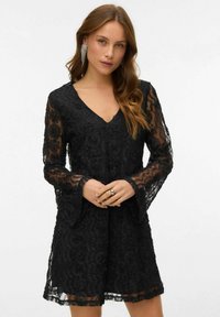 Černé krajkové šaty s výstřihem do V, dlouhými zvonovými rukávy, květinovými vzory a poloprůhlednou strukturou. Doplněno o prsten a dlouhé náušnice.