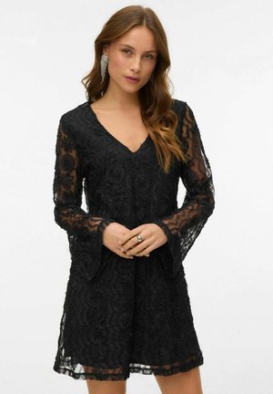 Crna haljina od čipke s V-izrezom, dugim rukavima u obliku zvona, cvjetnim uzorcima i poluprozirnom teksturom. Učinjena primjerena kombinacija s prstenom i dugim naušnicama.