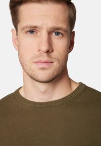 Oliwkowy sweter z dzianiny z okrągłym dekoltem, prążkowanymi mankietami i fakturą na ciele, noszony przez mężczyznę z krótkimi włosami i jasną karnacją.