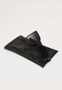 Ochelari de soare negri cu un design modern, unghiular, având lentile întunecate, așezați pe un toc din piele neagră, embossat cu "QUAY."