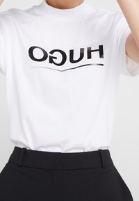 Camiseta de algodón blanca con el logo negro "HUGO" y detalle de línea curva, con un cuello alto y mangas cortas.