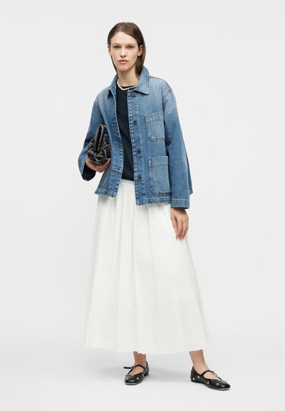 Γυναίκα που φοράει ένα τζιν μπουφάν, μαύρο τοπ, λευκή πλισέ φούστα, μαύρα παπούτσια με επίπεδη σόλα, περιδέραιο με μαργαριτάρια και κρατάει μια μαύρη τσάντα χειρός.