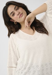 Lichtcrème blouse met een V-hals en lange mouwen, voorzien van gaatjespatronen en een textuurdesign. Subtiele accenten en details door het geheel.