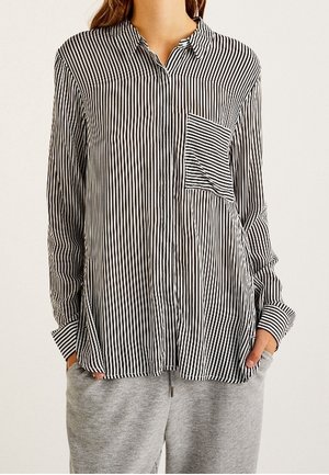 Frau trägt ein schwarz-weiß vertikal gestreiftes Hemd mit Knöpfen, einer kontrastierenden gestreiften Brusttasche und grauer Hose, die Hände in den Taschen.