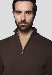 Brązowy sweter na zamek błyskawiczny z prążkowaną strukturą i stójką, z metalowym zamkiem, noszony na beżowej koszuli. Model prezentuje neutralny wyraz twarzy.