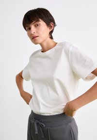 Hvid T-shirt med korte ærmer og rund halsudskæring, parret med grå bukser med elastisk talje. Stofdetaljen ser glat og åndbar ud.