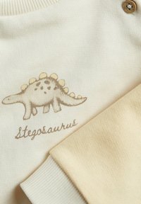 Vêtement en tricot beige avec un design de stégosaure en marron clair et des accents jaunes. Comprend un poignet beige et un détail de bouton en bois.
