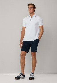 Polo bianco con colletto blu navy e logo "HACKETT", abbinato a pantaloni corti blu scuro. L'outfit è completato da sneaker nere e bianche.