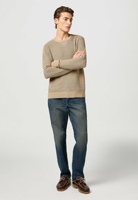 Maglione beige lavorato a maglia con colletto tondo, polsini e orlo a coste; abbinato a jeans blu e scarpe da barca marroni con lacci.