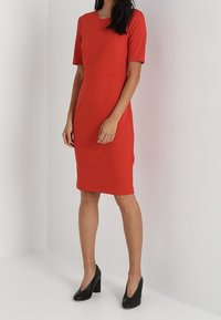 Czerwona dopasowana sukienka z krótkimi rękawami, o wyraźnej strukturze, do kolan, wykonana z gładkiego materiału. Noszona z czarnymi szpilkami.