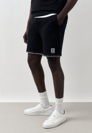 Férfi fekete rövidnadrágban, fehér varrással és kis virág logóval, fehér zoknival és fehér tornacipővel, zsebre tett kézzel áll.