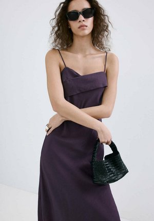 Mujer con cabello rizado, que lleva gafas de sol negras y un vestido sin mangas de color púrpura oscuro, sosteniendo un pequeño bolso negro tejido.