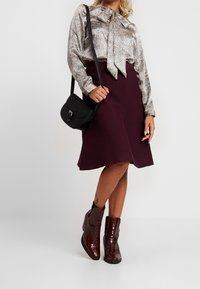 Blusa argento metallico con fiocco, gonna svasata bordeaux e stivaletti lucidi bordeaux. Piccola borsa a tracolla nera con dettaglio a chiusura.