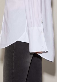 Chemise blanche à manches longues avec un ourlet fendu et des poignets à boutons, portée avec un jean gris. Le tissu semble lisse et légèrement texturé.
